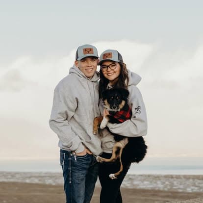 Couple in hoodies and hats holding a dog by the beach at sunset.