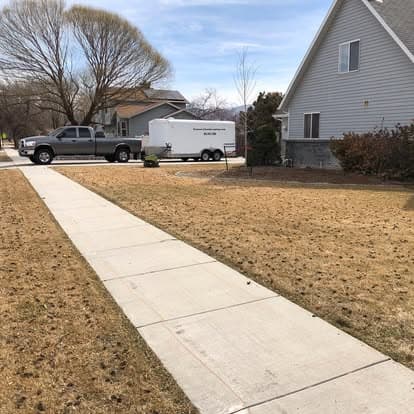 Front yard with dry grass, sidewalk, truck, and a parked trailer under a clear sky.