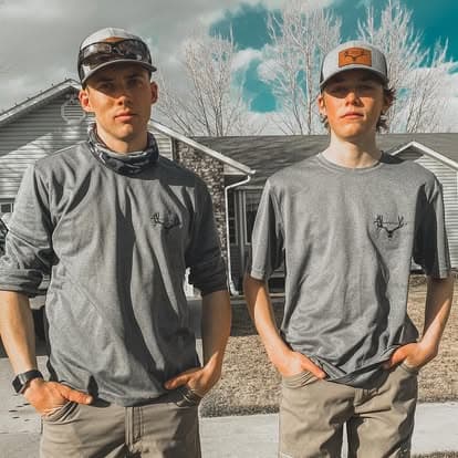 Two young men in matching gray shirts with antler logos, standing outdoors near a house.